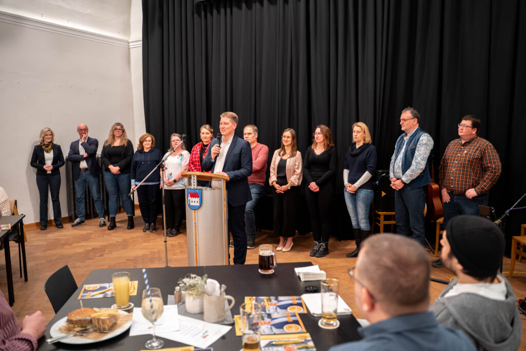 Foto der Kandidatinnen und Kandidatinnen der Liste für den Marktgemeinderat und Roland Vilsmaier am Rednerpult mit Mikrofon.